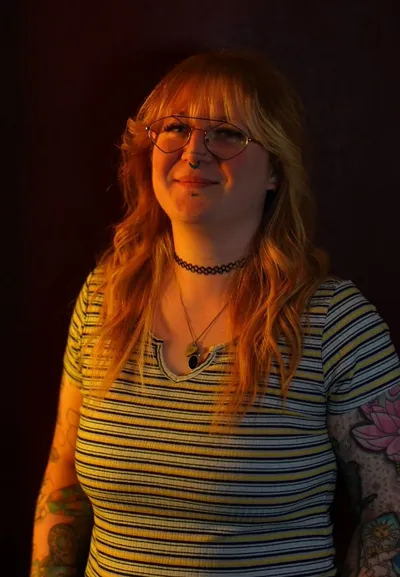 Kim, resident tattoo artist at Requiem Tattoo Shropshire, with strawberry blonde hair, hexagonal glasses and facial piercings, arms covered in colourful tattoos, lit by warm orange studio lighting against a dark background